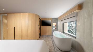 A modern bathroom with a freestanding bathtub, wooden paneling, a flat-screen TV, and a large window offering a city view.