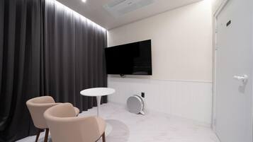 A modern room with a large flat-screen TV mounted on the wall, a round table, two chairs, and a door with a number.