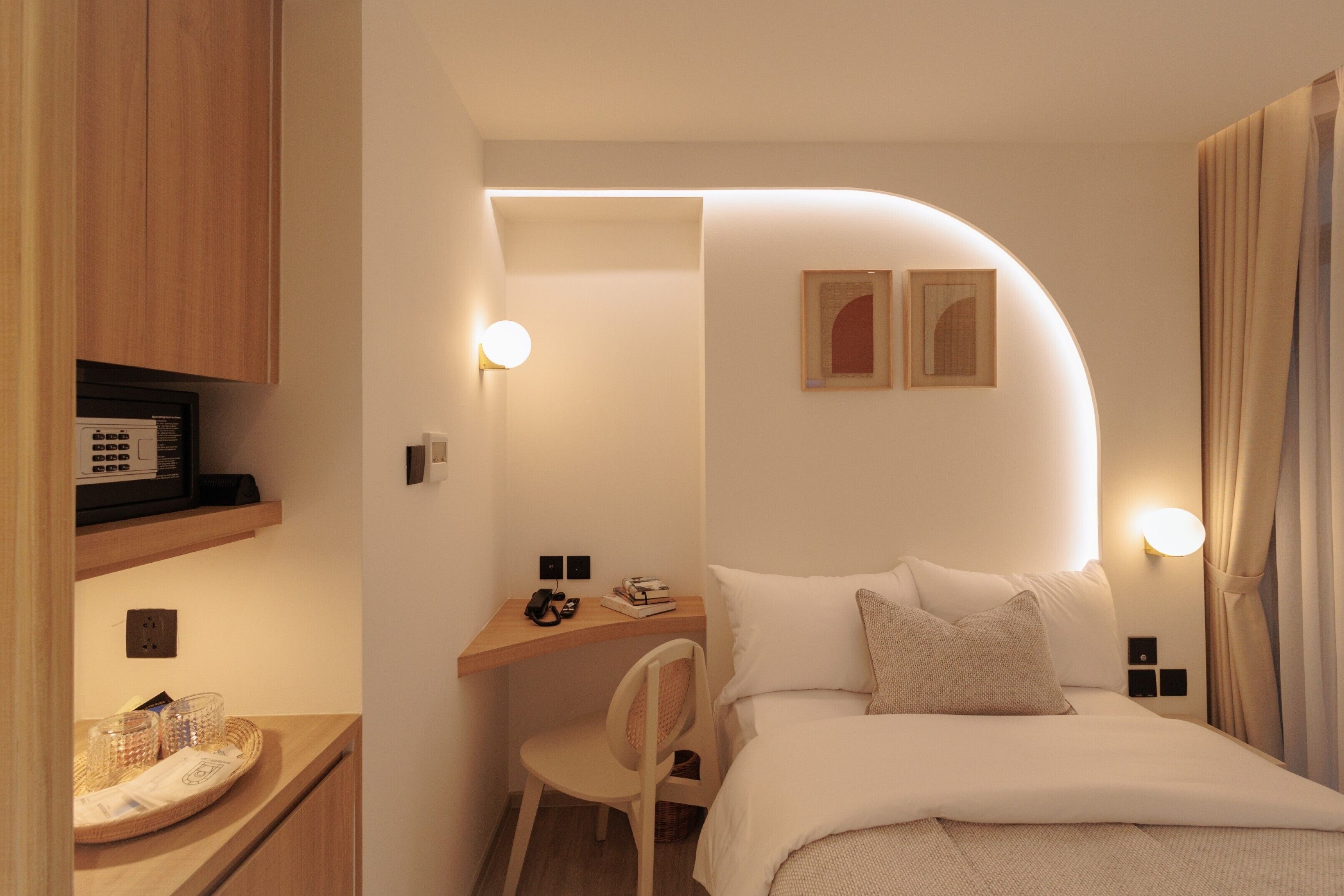 Habitación de hotel compacta con cama, escritorio y silla. Cuenta con dos lámparas fijadas a la pared y un estante pequeño con una radio y vasos.