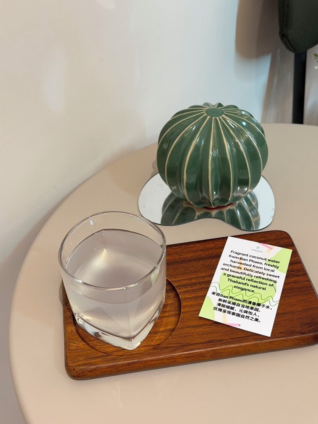 A glass of water on a wooden tray with a placemat, a decorative green object, and a card with text.