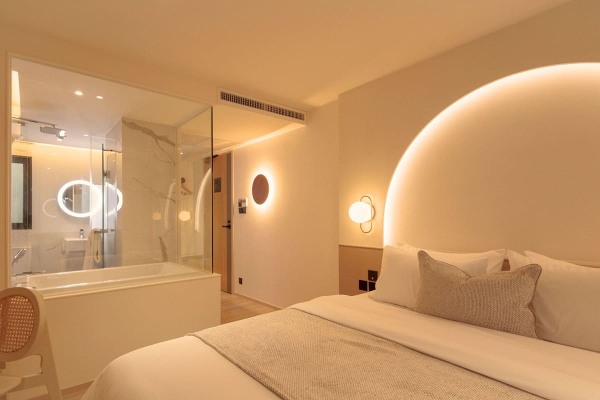 A modern hotel room with a large bed, a bathroom with a glass shower, and a mirror with lights.