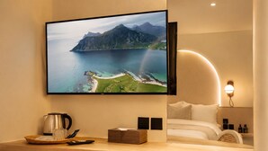 A hotel room with a large TV mounted on the wall, a wooden desk with a kettle, a remote control, and a basket, a bed with white bedding, and a wall-mounted light fixture. - Corridor2407 Hotel (Bangkok)