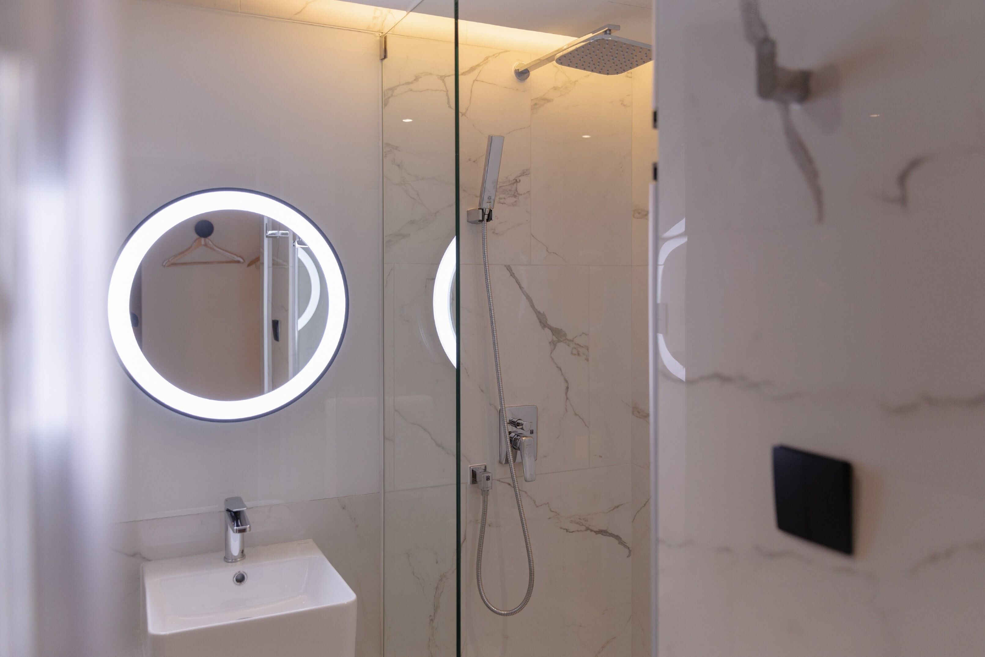 Un baño moderno con ducha de vidrio, un espejo redondo con luz y un lavamanos blanco.