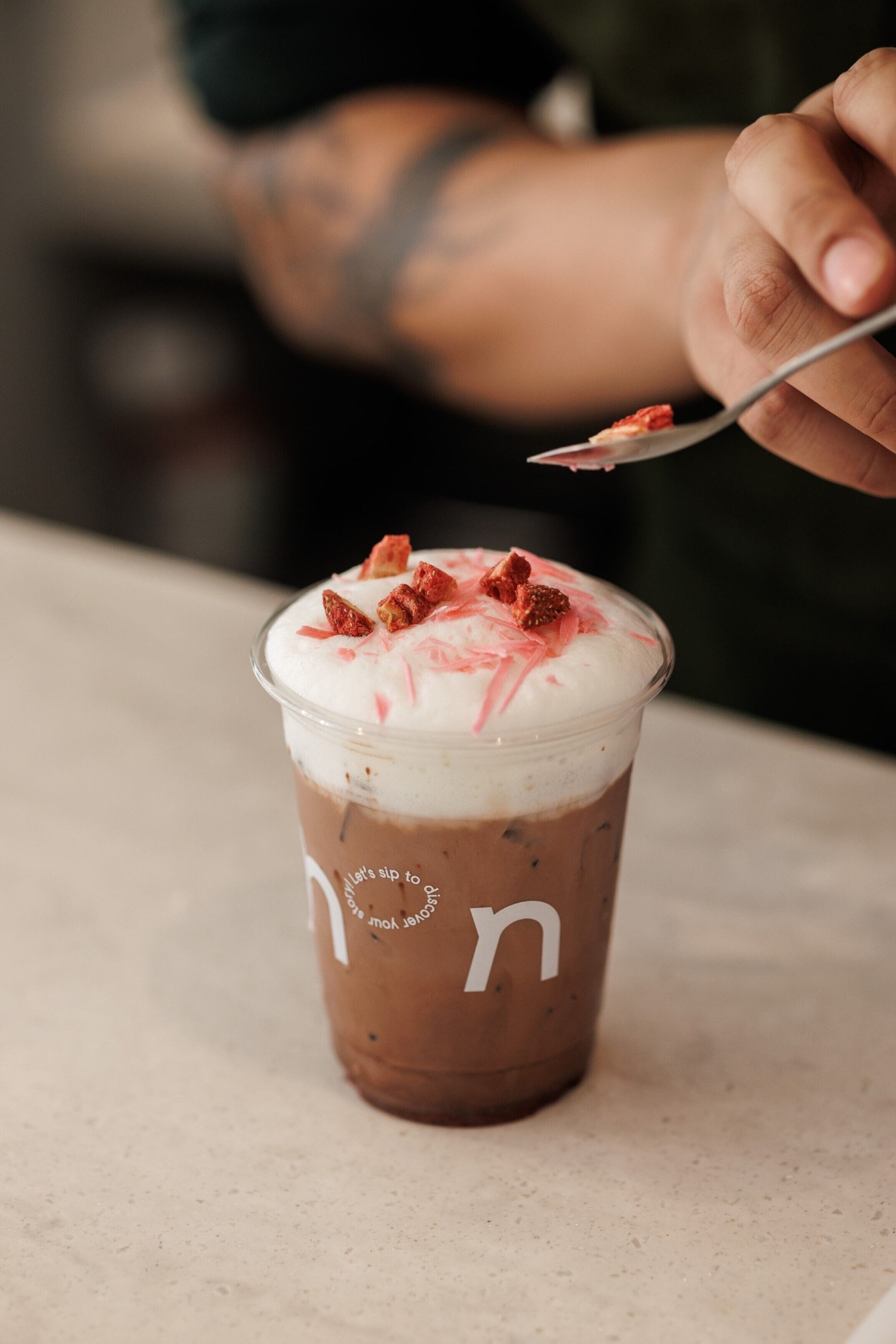 A clear plastic cup with a lid, containing a layered beverage with a whipped topping and a spoon inside.