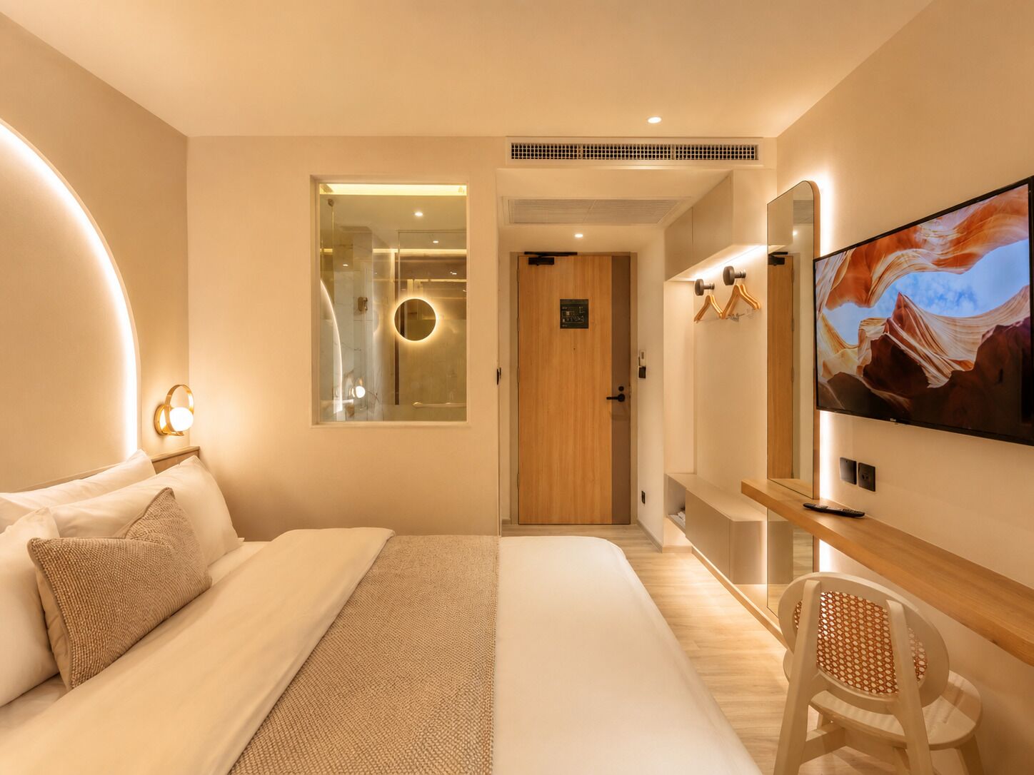 A modern hotel room with a bed, a television, a desk, and a bathroom visible through a glass door.