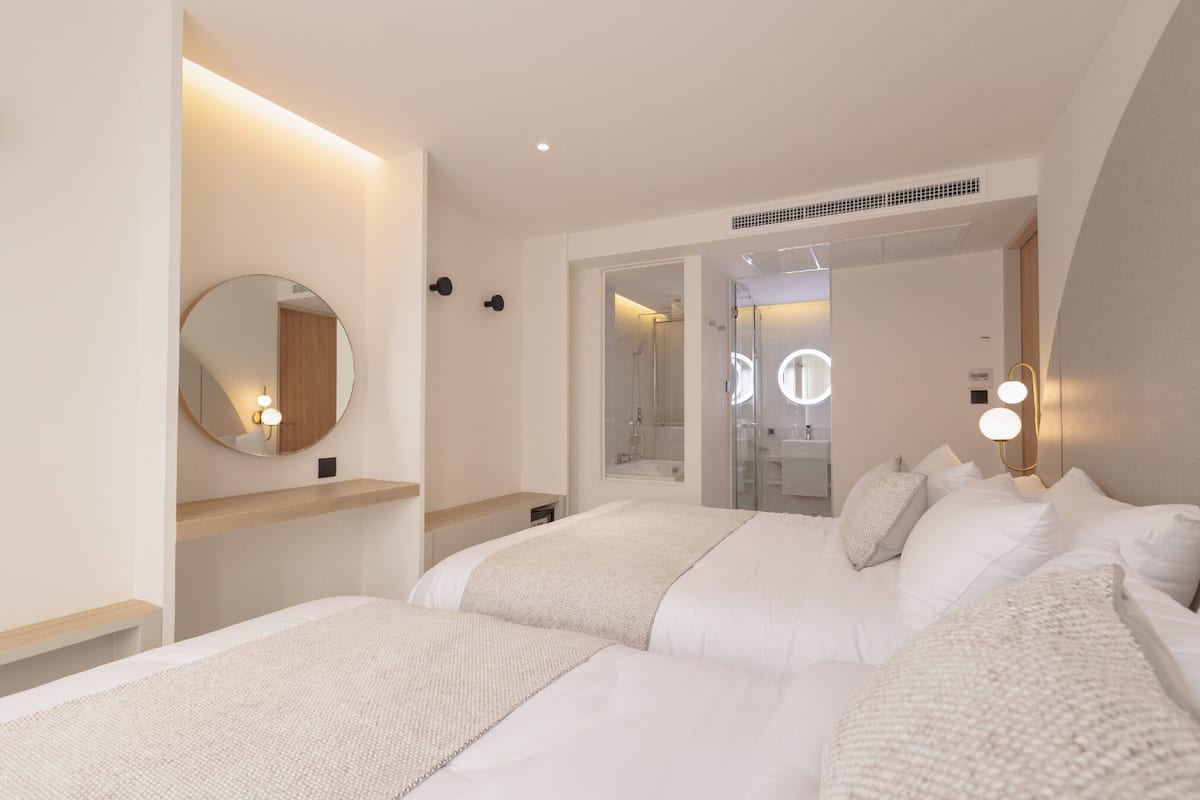 A modern hotel room with two beds, a round mirror, and a bathroom visible through an open door.
