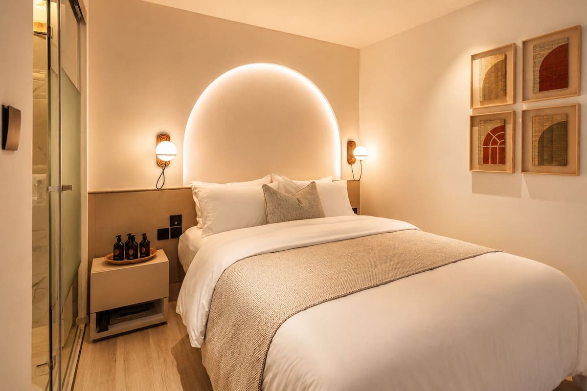 A modern hotel room with a large bed, bedside tables, wall-mounted artwork, and a glass shower enclosure.