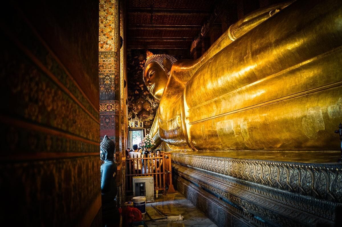A large golden Buddha statue in a temple with intricate wall carvings and a small shrine.