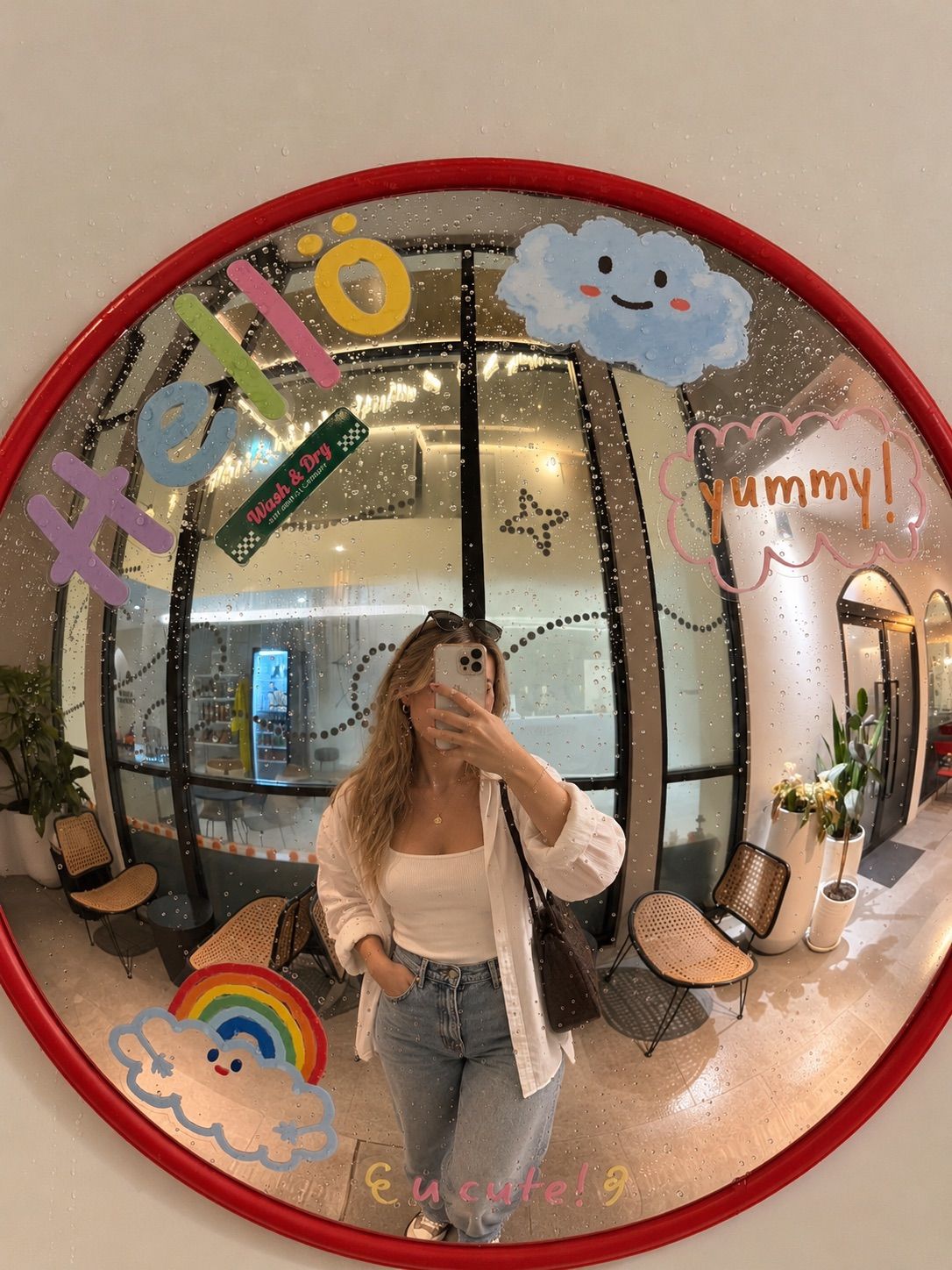 A person taking a selfie in a room with a large mirror reflecting a cafe interior, a rainbow sticker, and a cloud sticker.