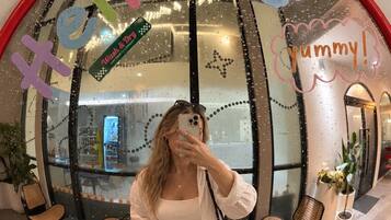 A person taking a selfie in a room with a large mirror reflecting a cafe interior, a rainbow sticker, and a cloud sticker.