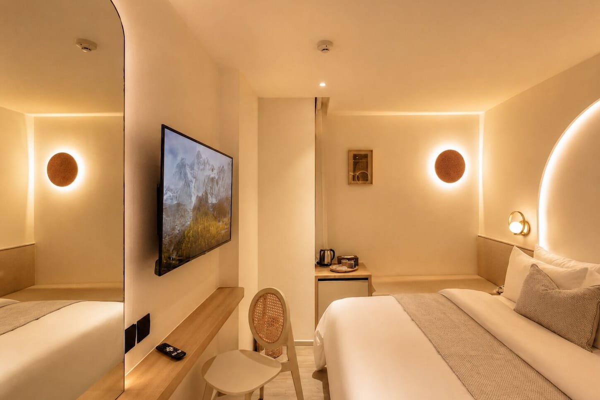 A hotel room with two beds, a TV mounted on the wall, a chair, a small table with a kettle, and a framed picture on the wall.