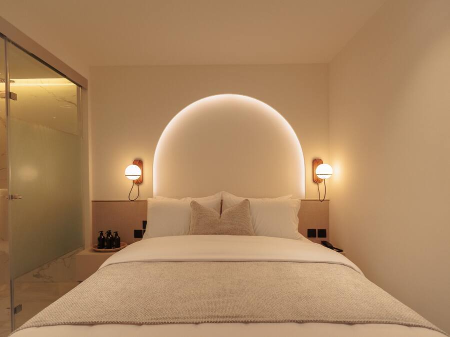 A modern hotel room with a large bed, two bedside tables, a glass shower enclosure, and wall-mounted lighting.