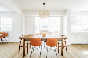 Un comedor luminoso con una mesa de madera y sillas naranja, una lámpara colgante, y vistas a una cocina y sala de estar a través de amplios ventanales.