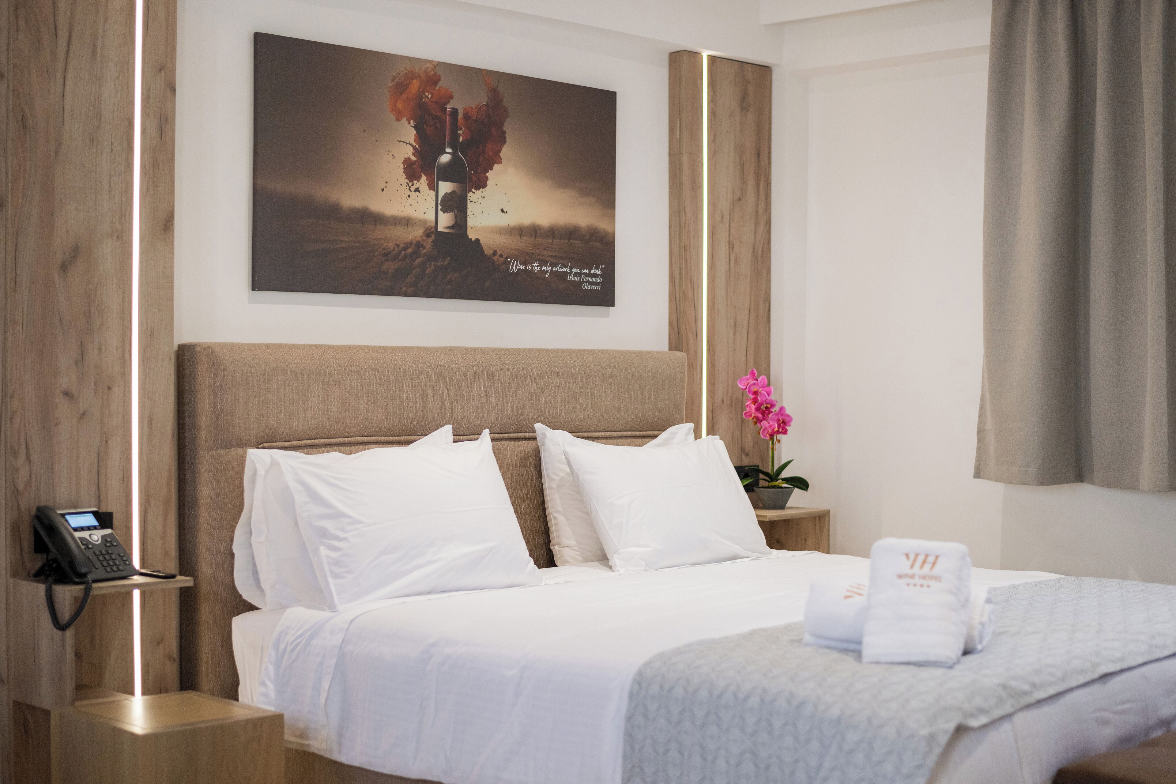 A hotel room with a large bed, white bedding, a mounted artwork, a bedside phone, and a small table with a pink flower.