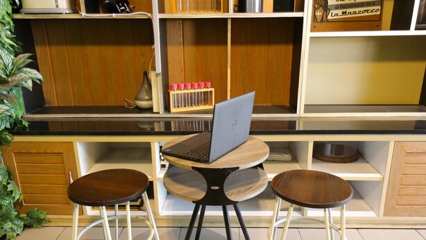 Una cocina con una laptop sobre una mesa de madera, dos taburetes y una alacena con utensilios de cocina.