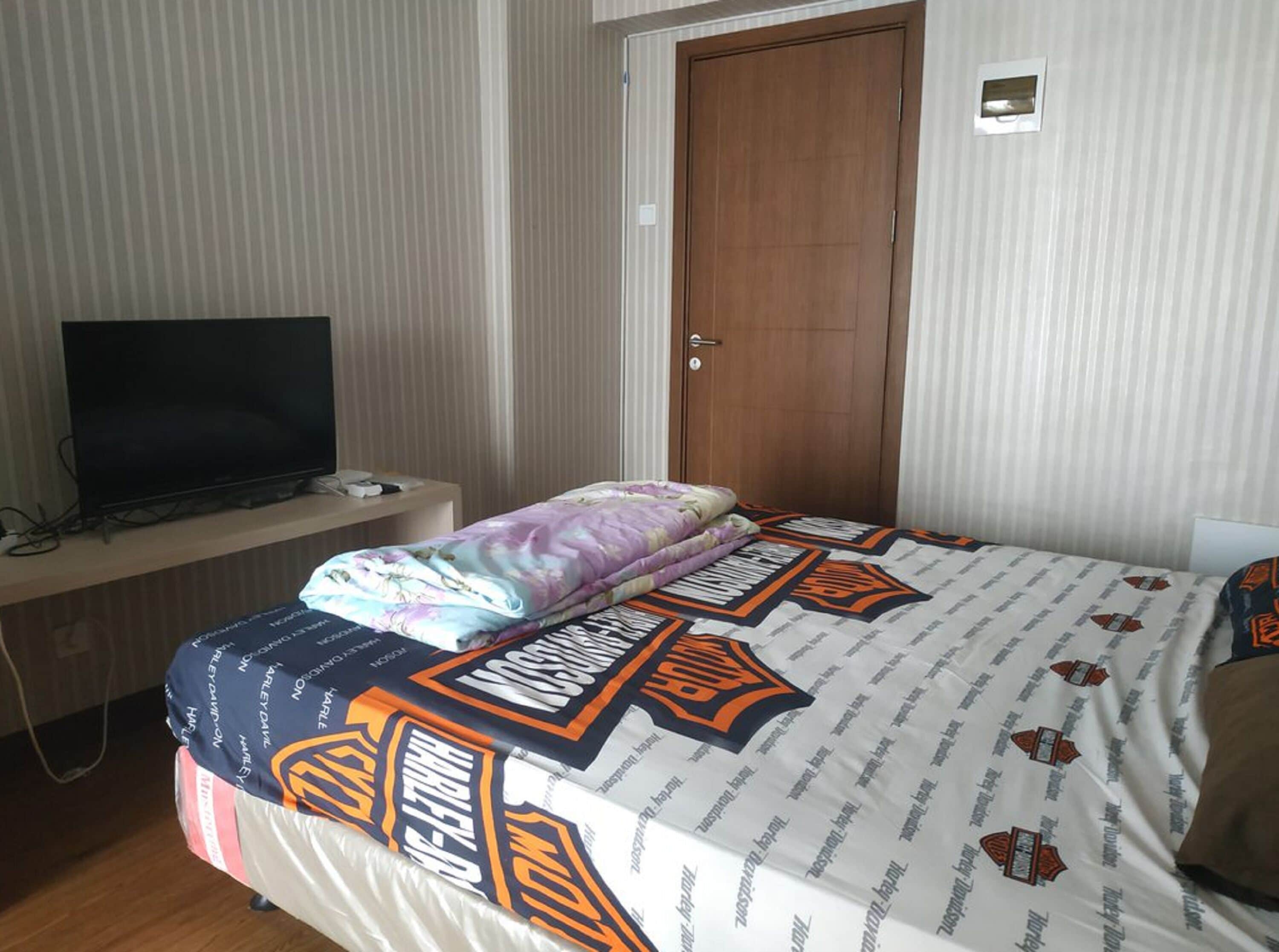 A hotel room with a bed, a TV on a desk, and a door.
