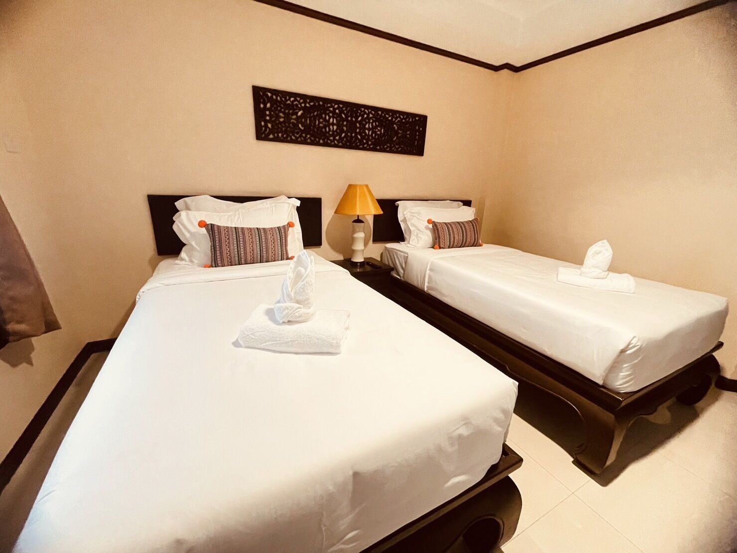 A hotel room with two single beds, each with white linens and pillows, a bedside table with a lamp, and a decorative wall piece above the beds.