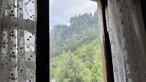 Une vue à travers une fenêtre ornée de rideaux à motifs floraux, dévoilant un paysage montagneux verdoyant, parsemé de quelques bâtiments.