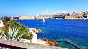 Un paisaje urbano costero con mar azul cristalino, un velero y un muro fortificado al fondo.