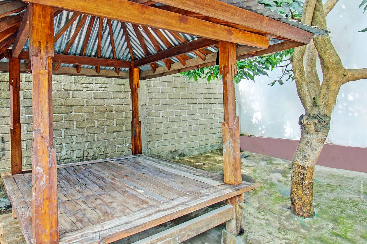 A wooden pergola structure with a bench underneath, set against a brick wall and a tree.