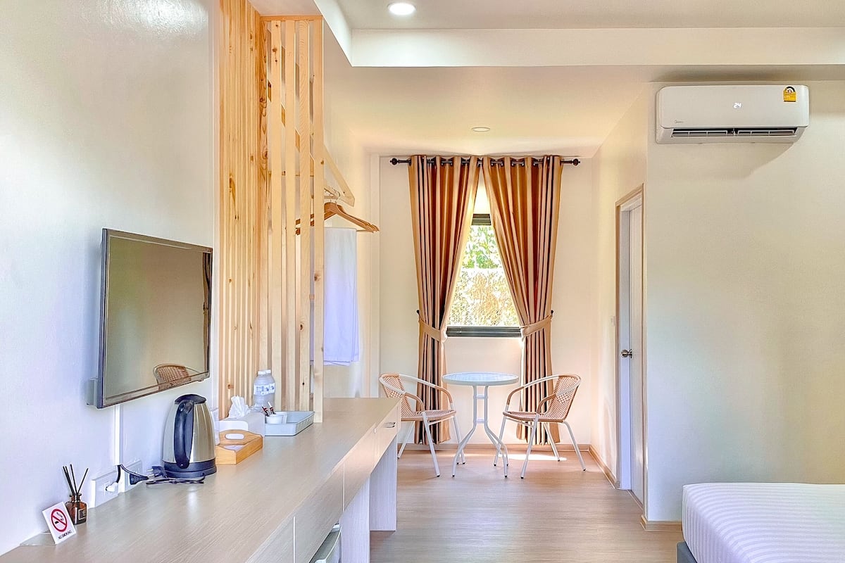 A hotel room with a bed, a TV, a small table with chairs, a window with curtains, and an air conditioning unit.