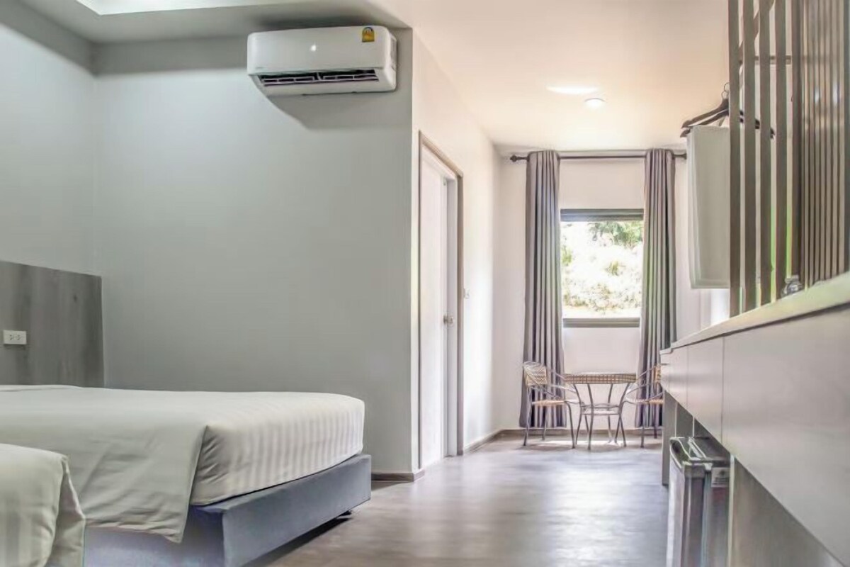 A hotel room with a bed, a small table with chairs, an air conditioning unit, and a view of the outdoors through a window.
