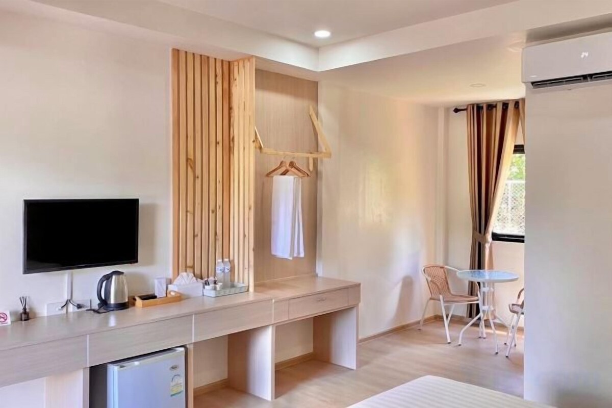 A modern hotel room with a flat-screen TV, a wooden desk, a mini-fridge, a hanging coat rack, and a small dining area with a table and chairs.