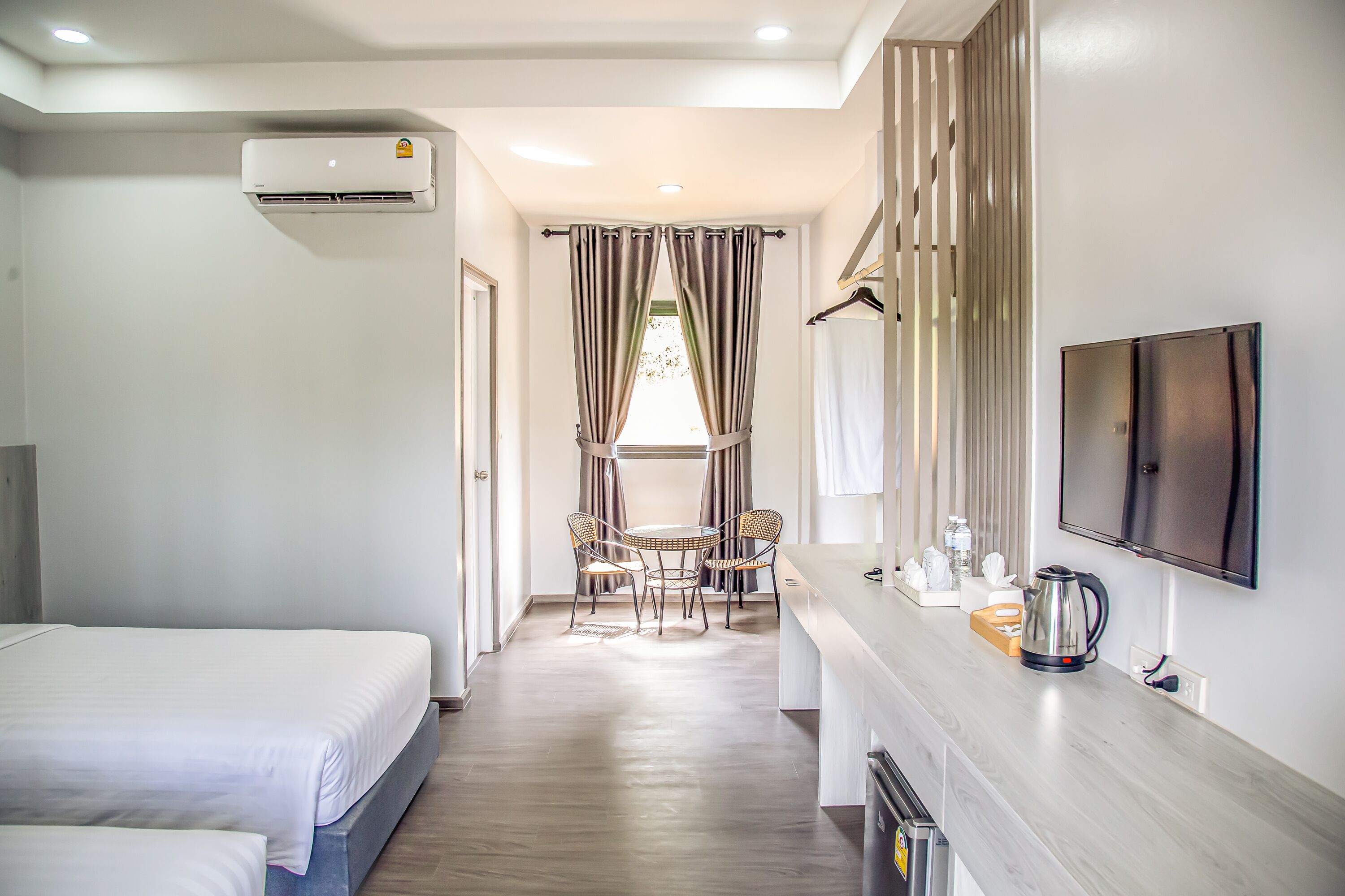 A hotel room with two beds, a dining area with a table and chairs, a kitchen counter with a kettle, and a TV mounted on the wall.