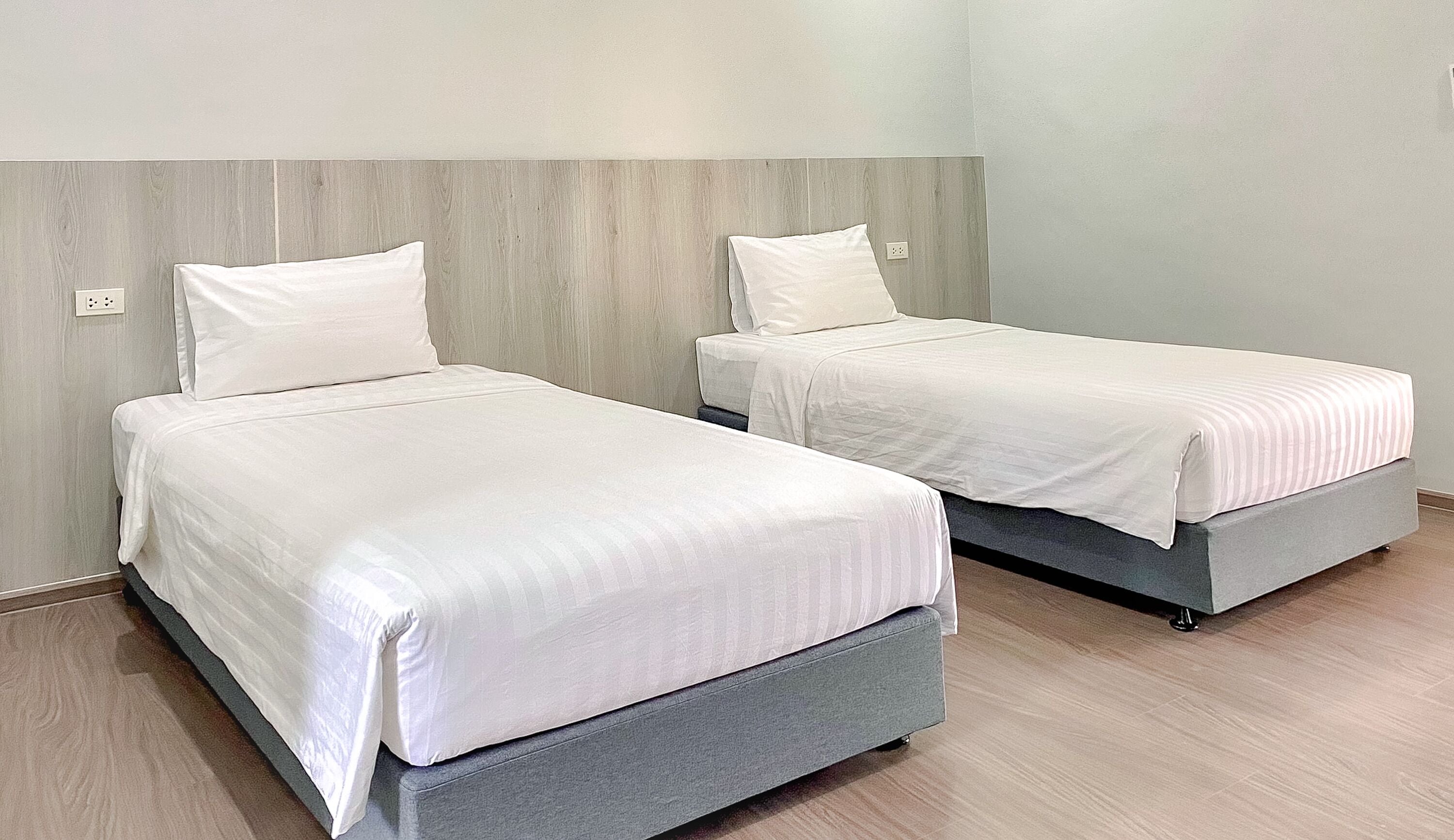 Dos camas individuales con bases grises y ropa de cama blanca en una habitación con paredes y suelo de madera clara.