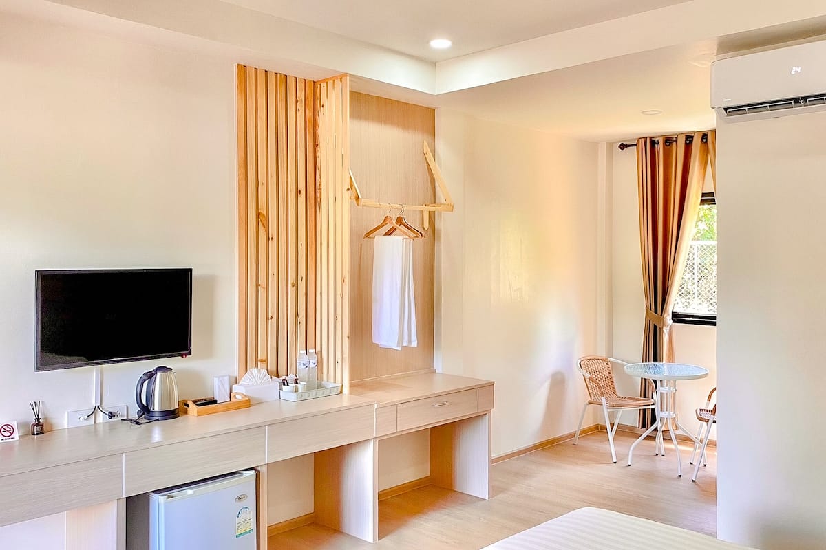 A modern hotel room with a wooden desk, a television, a mini-fridge, a small table with chairs, and a window with curtains.
