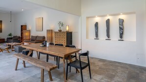Un vaste espace de vie comprenant une table à manger en bois, des chaises noires et un banc. On y trouve également un ensemble de canapés, une table basse et une bibliothèque.