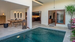 Une piscine intérieure moderne avec une terrasse en bois, un coin repas comprenant une table et des chaises, et une cuisine en arrière-plan.
