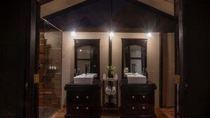 A modern bathroom with two dark wooden sinks, a glass shower enclosure, and a small shelf with toiletries. - Leopard Trails Yala (Kataragama)