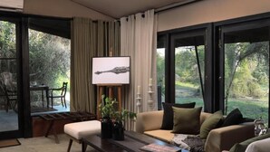 A modern living room with a sofa, coffee table, and a painting on the wall. There is a large window with curtains, and a view of greenery outside. - Leopard Trails Yala (Kataragama)