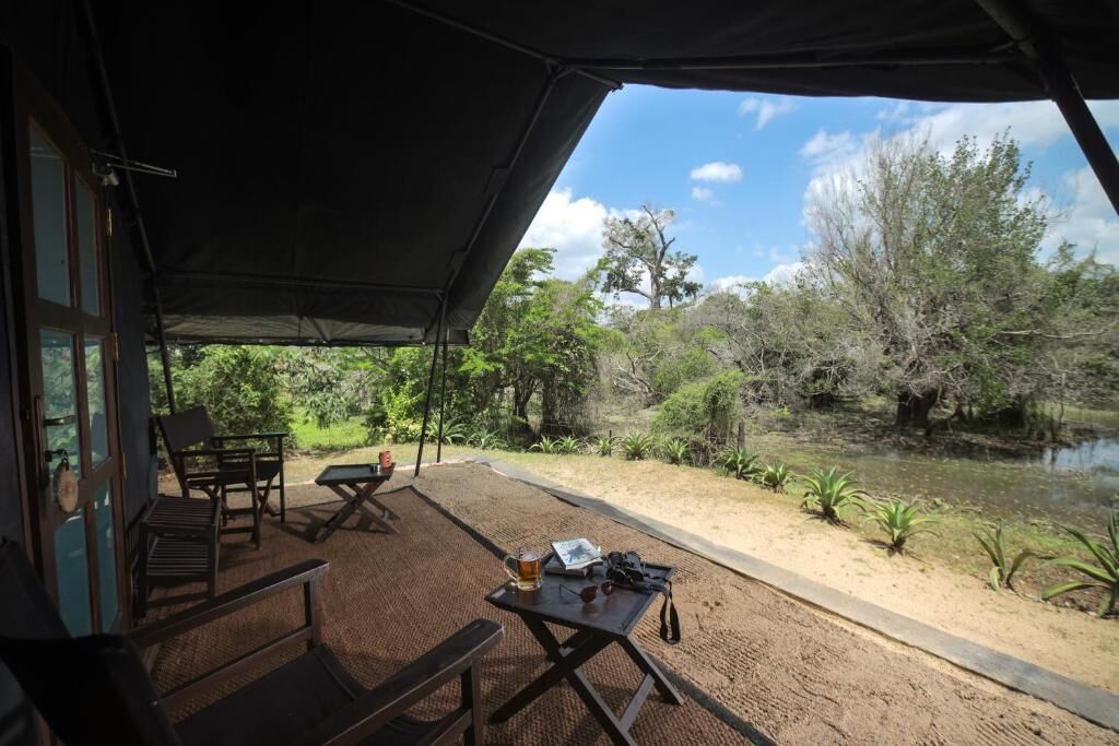 An outdoor seating area with tables and chairs, a covered section with a table and a chair, and a view of a natural landscape with trees and a water body.