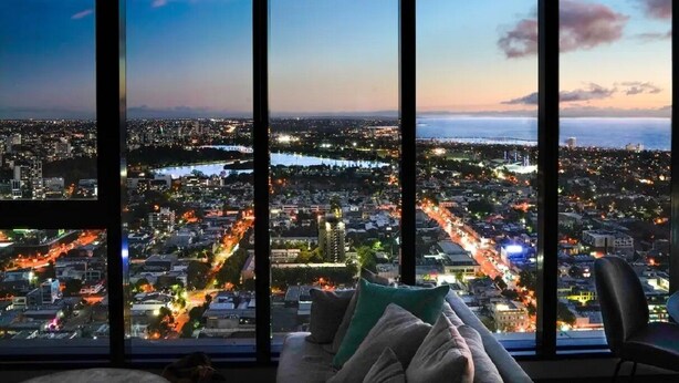 Ein Hotelzimmer in einem Hochhaus mit großem Fenster und Blick auf die Stadt bei Abenddämmerung.