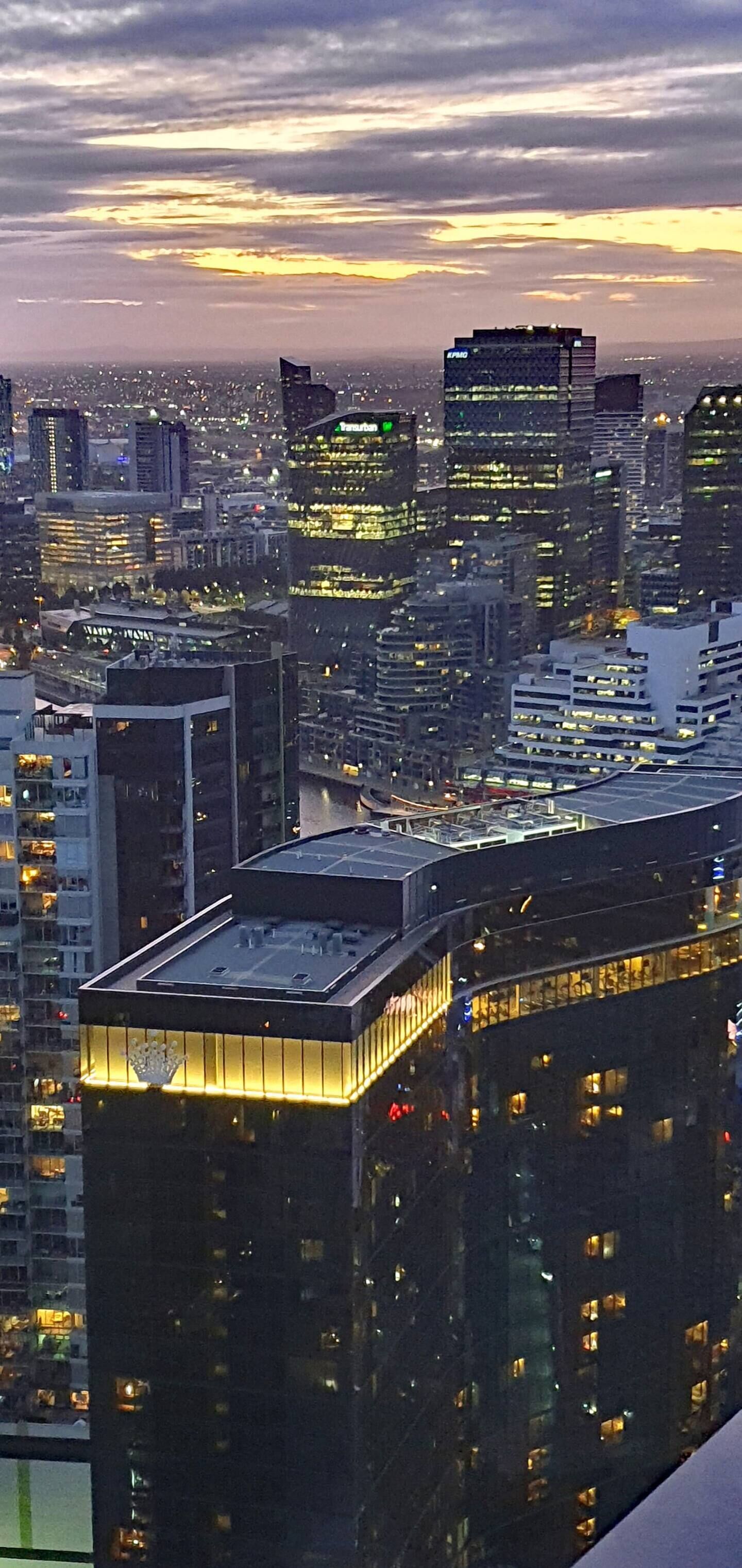 A cityscape at dusk with illuminated buildings and a river in the foreground.