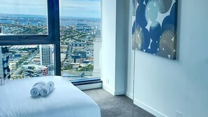 Ein Hotelzimmer mit großem Fenster und Blick auf die Stadt, einem ordentlich bezogenen Bett und einem modernen, abstrakten Gemälde an der Wand.