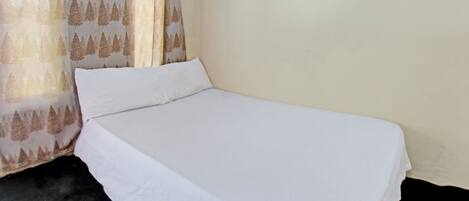 Una cama individual con ropa blanca en una habitación con pared lisa y cortina estampada con motivos de árboles.