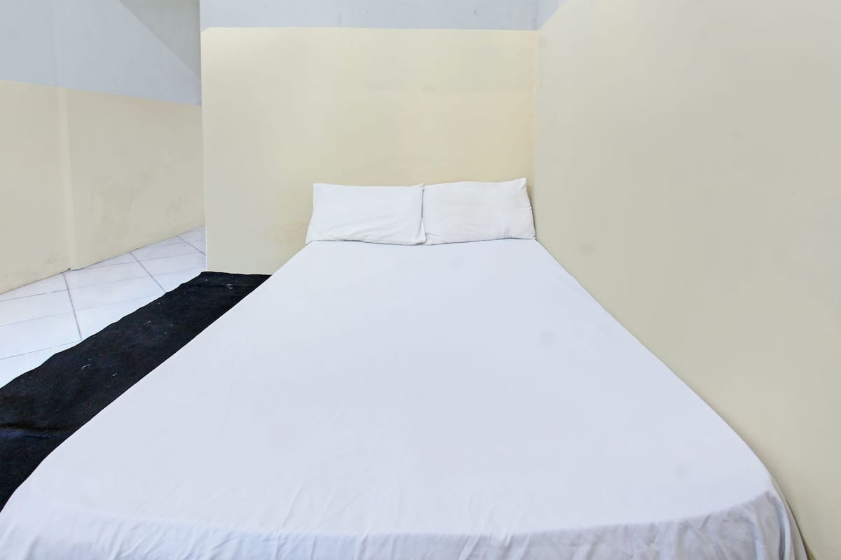 A small, single-bed room with a plain white bedspread and pillows, a black rug, and plain walls.