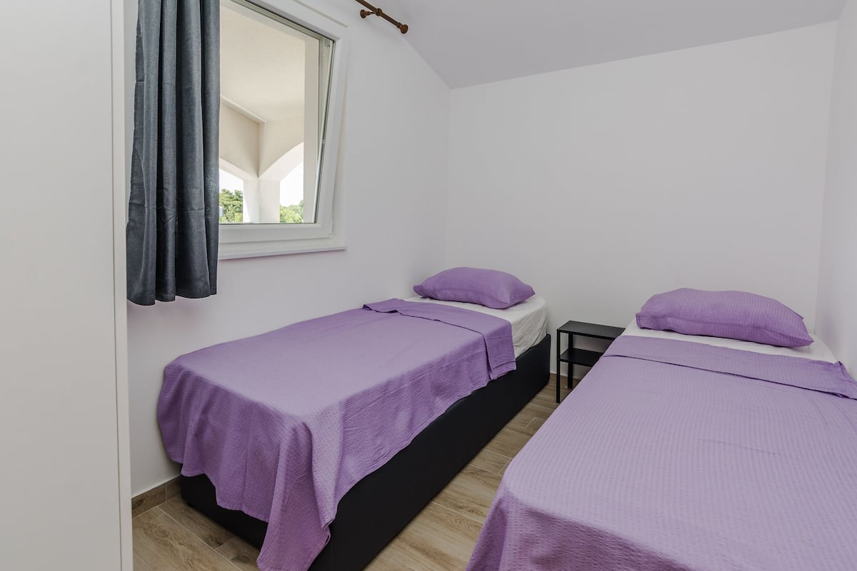 Une petite chambre propre, avec deux lits simples, une fenêtre avec des rideaux et un plancher en bois.