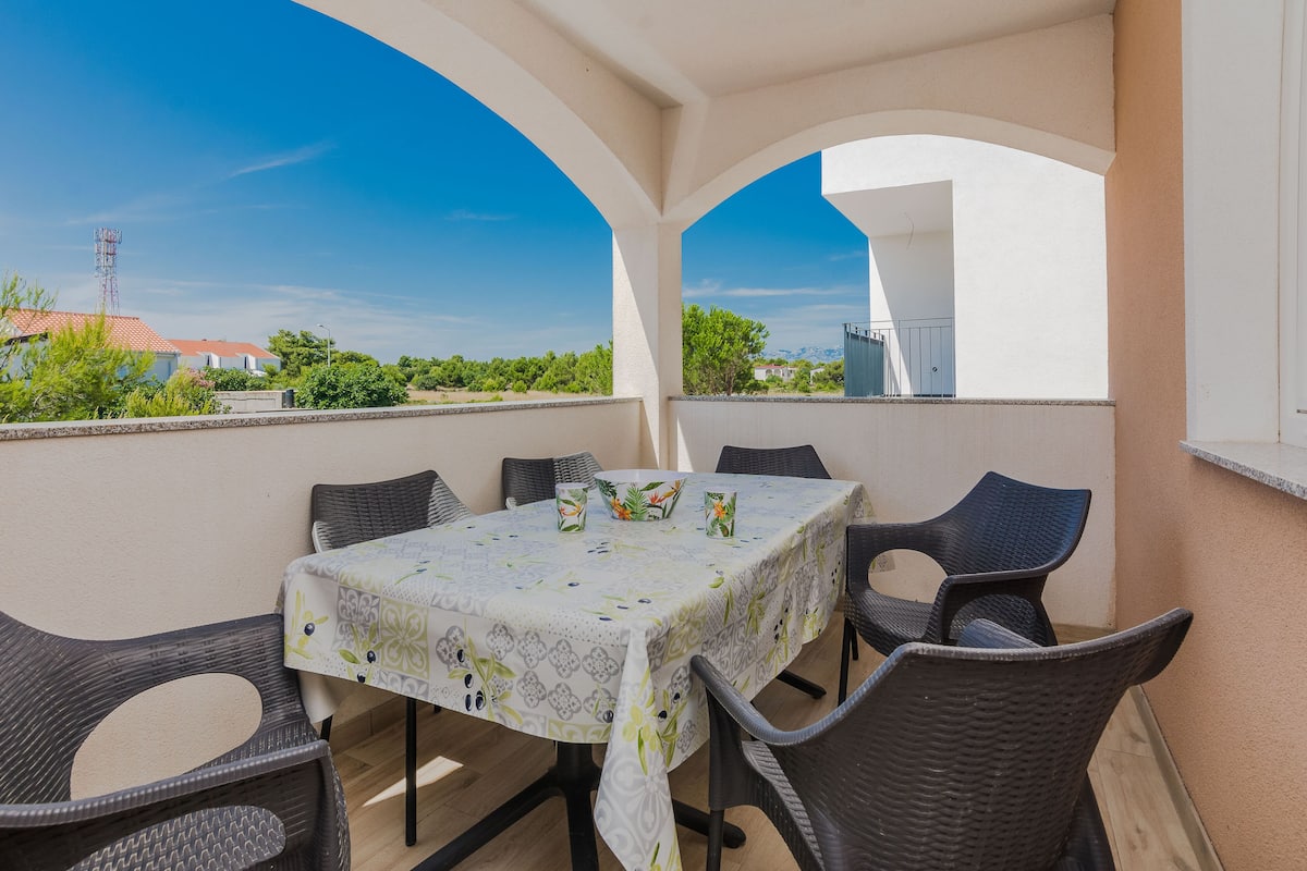 Une terrasse extérieure avec une table et des chaises, un ciel bleu dégagé et une vue sur la verdure et les bâtiments en arrière-plan.