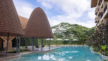 Una piscina con sillas de descanso y sombrillas, rodeada de follaje y un edificio con una estructura de techo cónica distintiva.