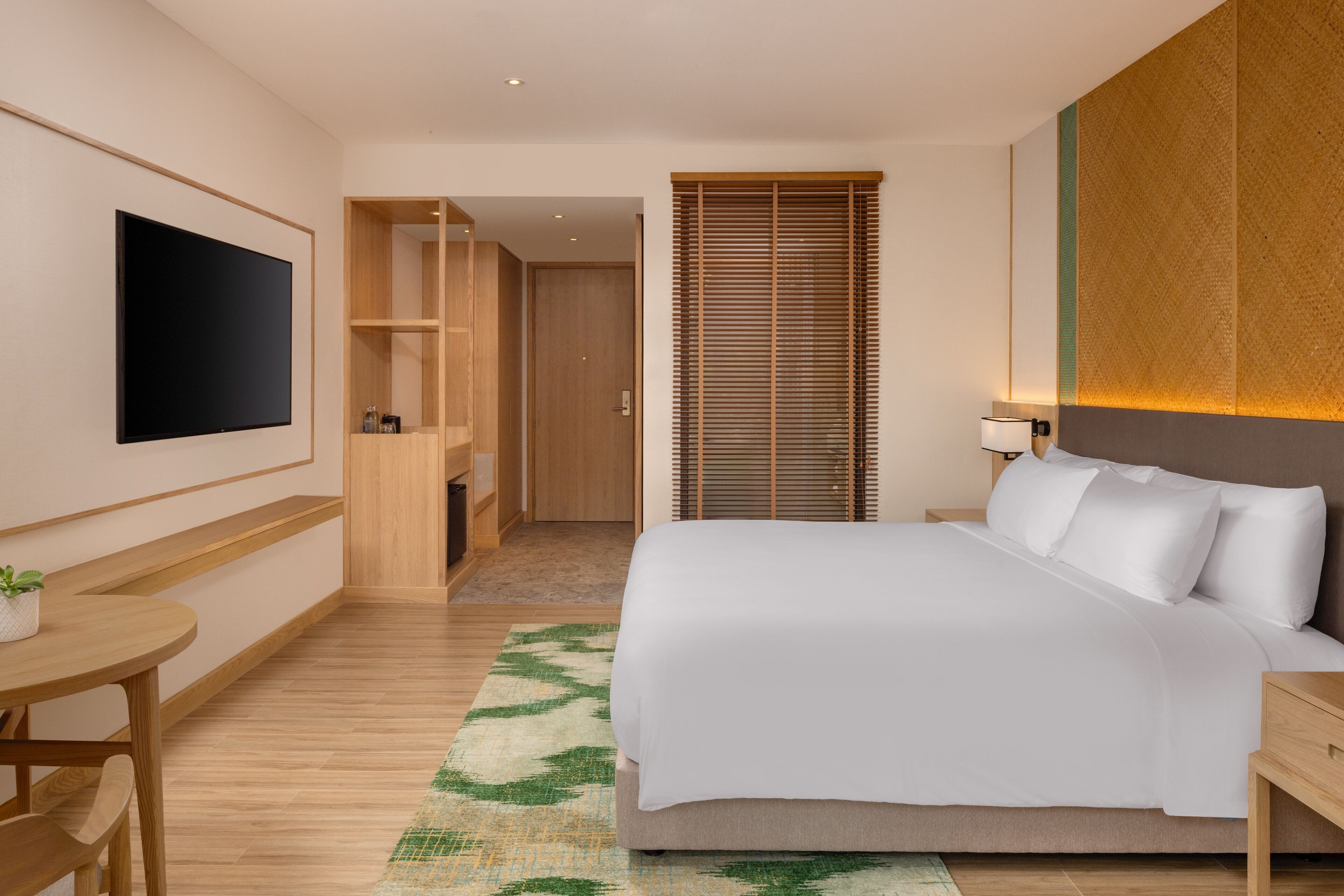 A modern hotel room with a large bed, a flat-screen TV mounted on the wall, a wooden desk, and a small table with a plant.
