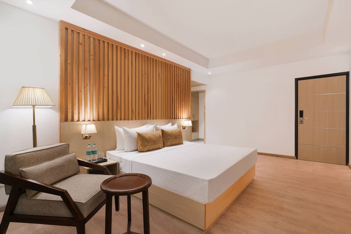 A modern hotel room with a large bed, a wooden headboard, a sitting area with a chair and table, and a nightstand with a lamp and water bottles.