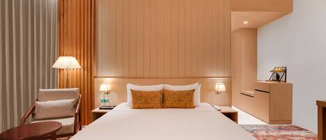 A hotel room with a large bed, a wooden headboard, a chair, a nightstand with a lamp, and a desk with a lamp.