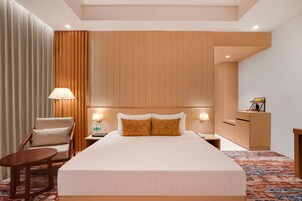 A hotel room with a large bed, a wooden headboard, a chair, a nightstand with a lamp, and a desk with a lamp.