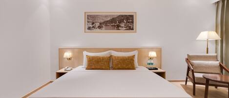 A hotel room with a large bed, a nightstand with a lamp, a chair, and a framed picture on the wall.