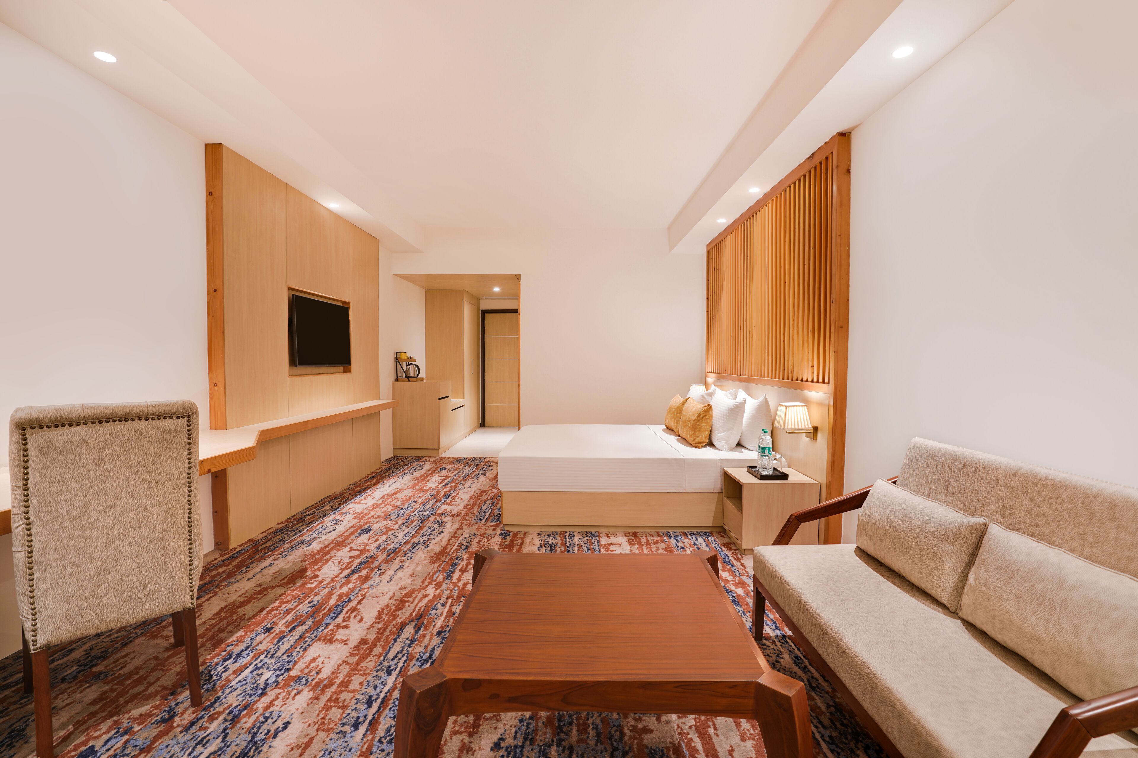 Una habitación de hotel con una cama, un sofá, una mesa de centro de madera, un televisor montado en la pared y una alfombra estampada.
