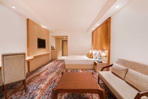 Una habitación de hotel con una cama, un sofá, una mesa de centro de madera, un televisor montado en la pared y una alfombra estampada.